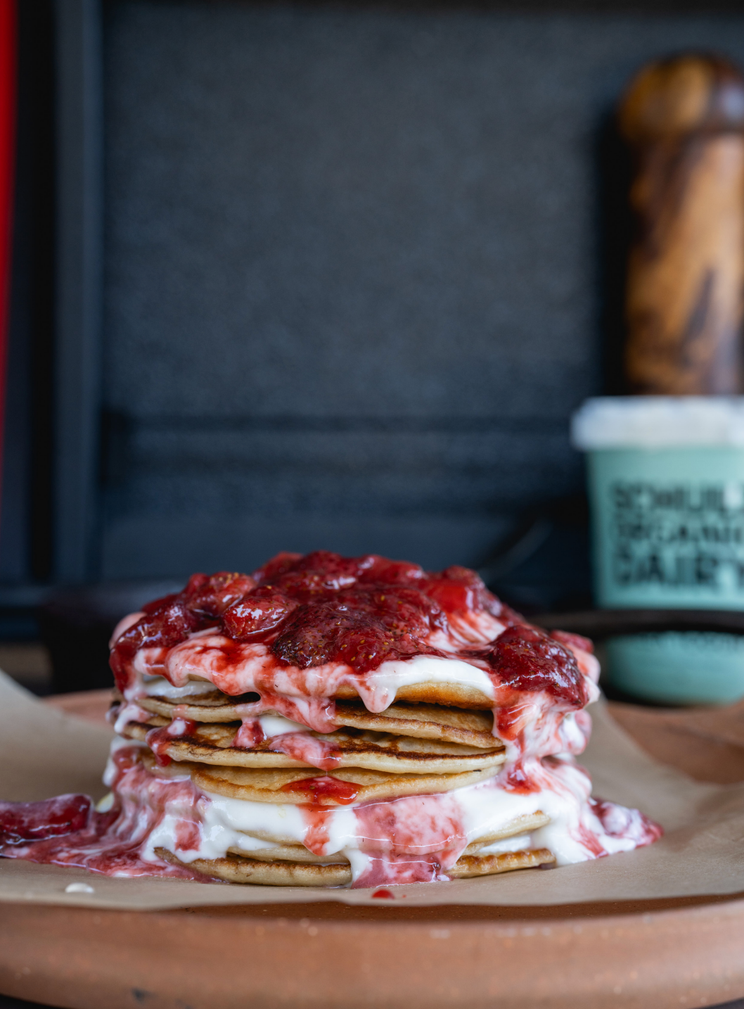 A close-up shot of a stack of pancakes topped with a thick layer of red berry sauce and white cream, sitting on parchment paper with a blurred background.